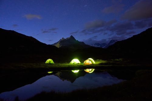 Free A tranquil night camping by a mountain lake with glowing tents under a starlit sky. Stock Photo
