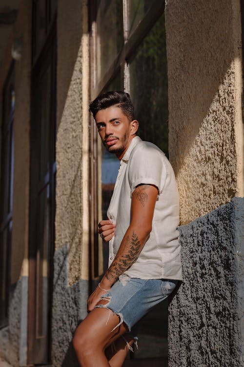 Man in White Shirt and Denim Shorts Posing by Wall