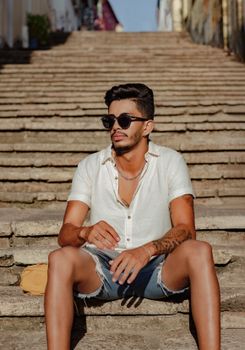A stylish young man in a summer outfit relaxing on an urban staircase in sunlight.