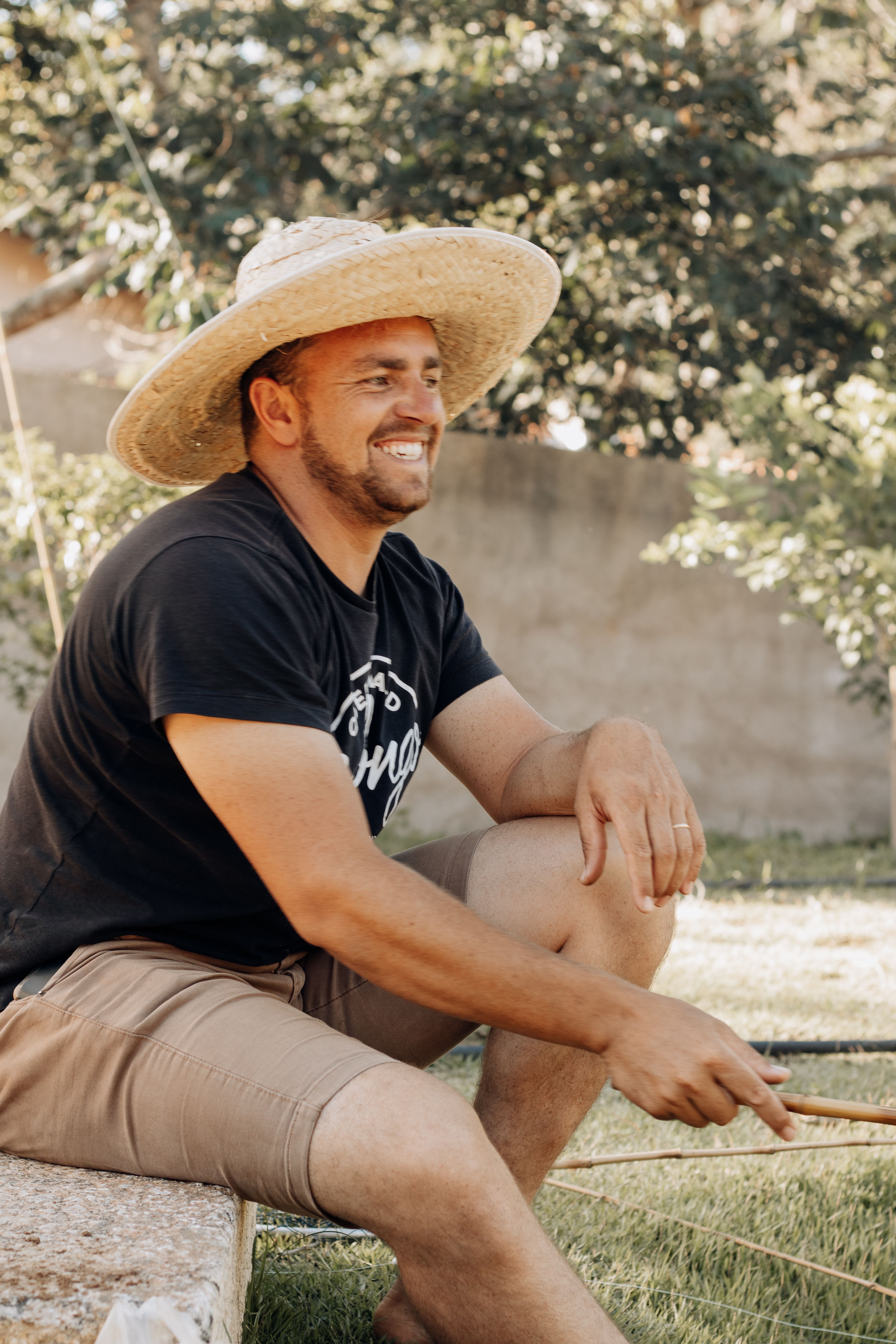 Casual portrait of a smiling man seated outdoors in São Paulo wearing a straw hat and black t-shirt.