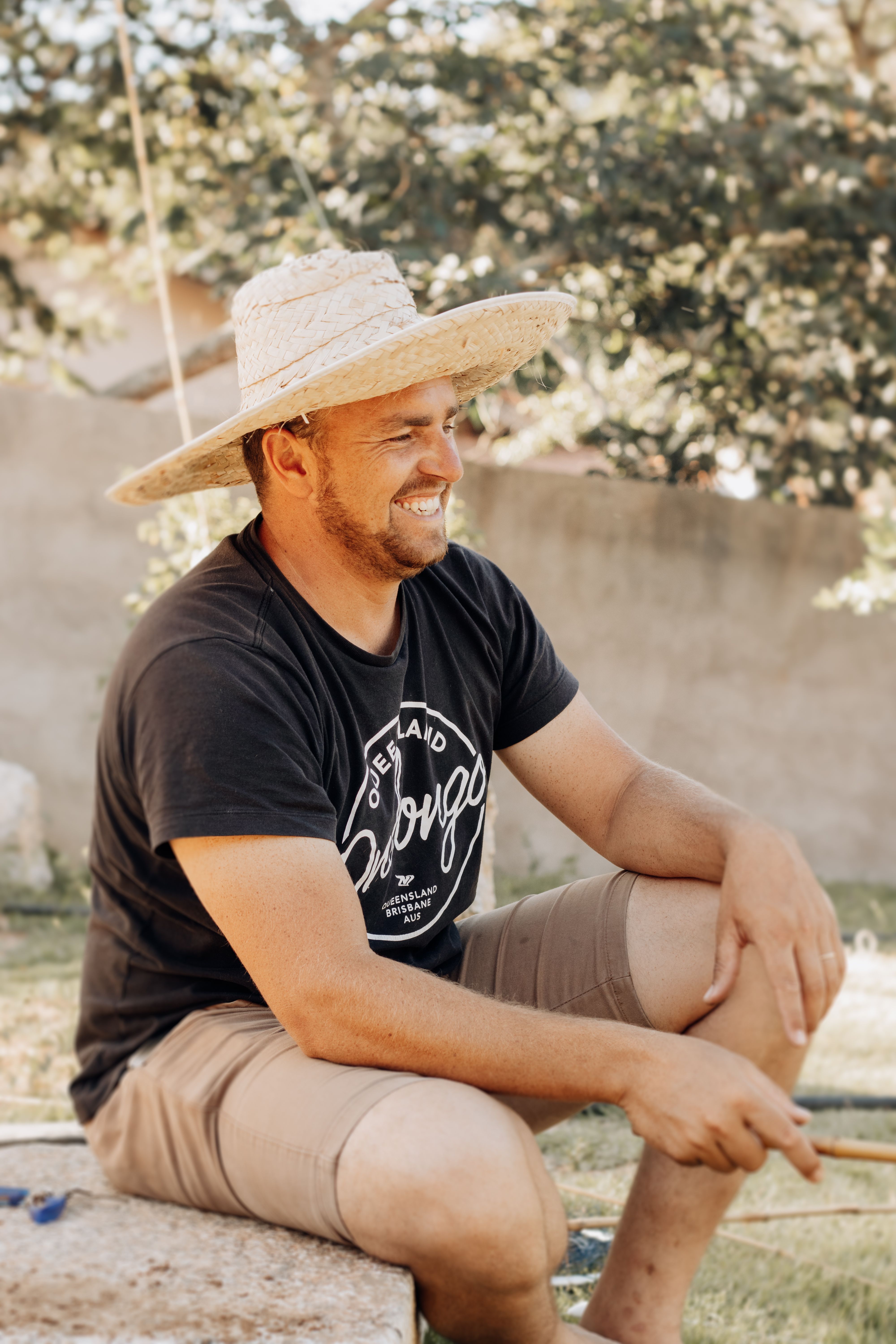 Casual outdoor portrait of a smiling man in São Paulo, wearing a black t-shirt and wicker hat, exuding relaxation.