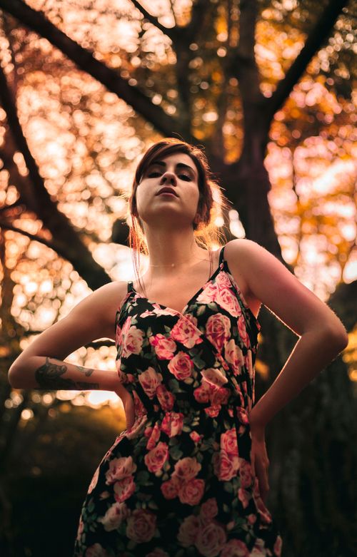 Free Charming woman in a floral dress poses confidently amongst trees during sunset. Stock Photo