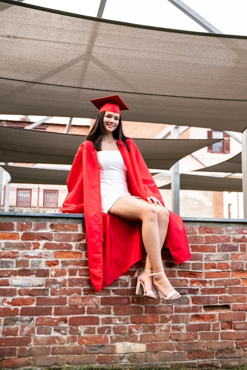 Smiling Graduate Sitting in Academic Hat and Gown on Wall