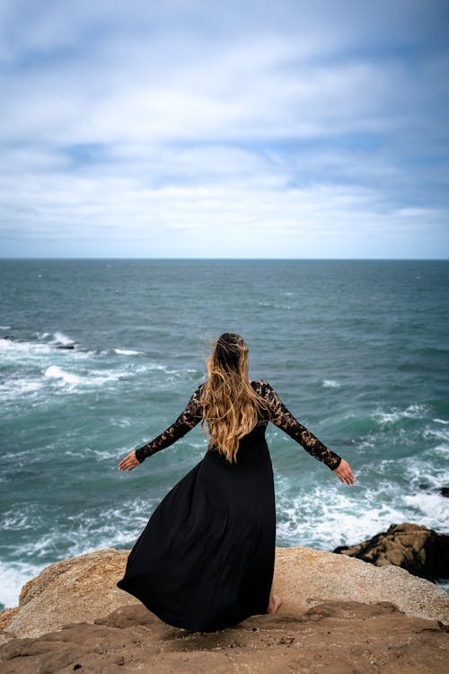 Back View of Woman in Black Dress Standing on Rocks on Sea Shore