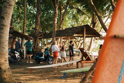 Group of people enjoying a tropical beach setting with motor scooters and surfboards under palm trees.