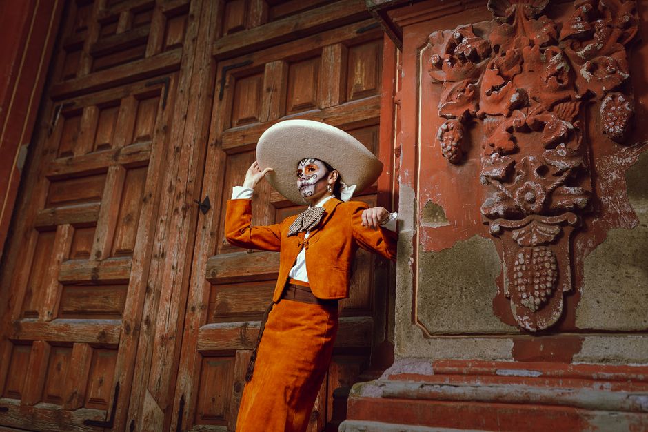 Person in traditional Mexican attire with face art stands by an ornate wooden door.
