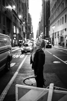 A woman looks up on a bustling New York City street at dusk, captured in black and white.