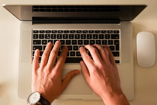 Hands typing on a laptop keyboard, showcasing a modern workplace environment with a mouse nearby.