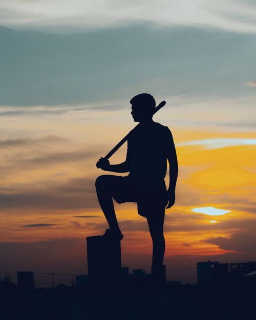 Free Sunset silhouette of a man holding a bat on a rooftop, capturing an urban skyline in India. Stock Photo