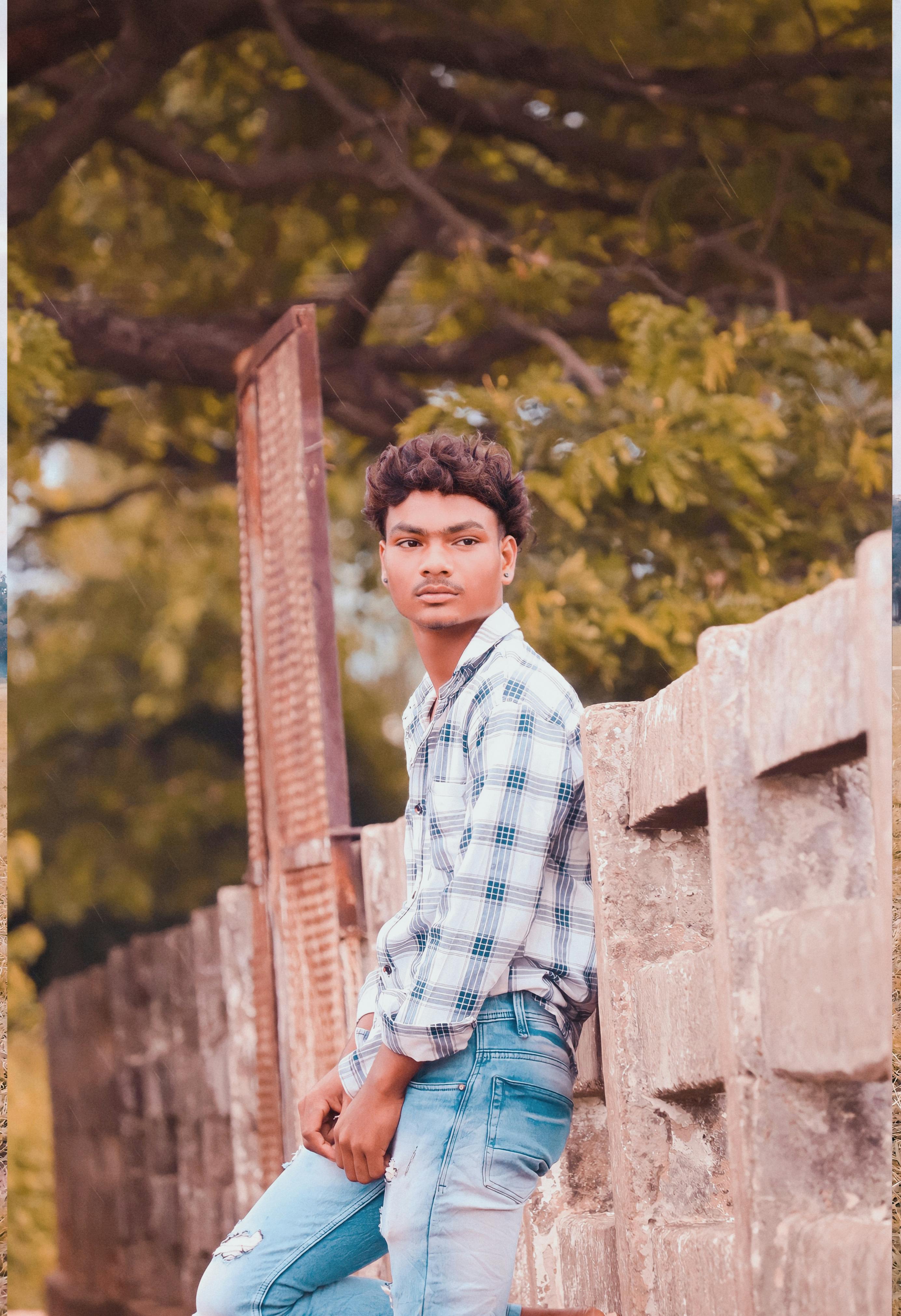 Portrait of a young man in a checked shirt and jeans leaning against a rustic concrete fence outdoors.
