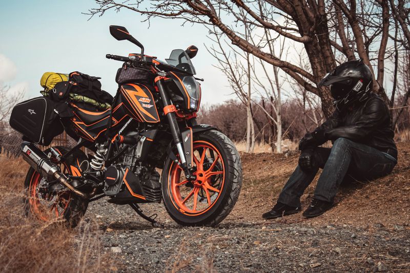 A motorcyclist in gear rests beside a packed KTM bike on a rural path