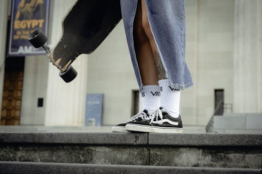 A stylish woman stands with a skateboard, wearing a denim skirt and black sneakers.