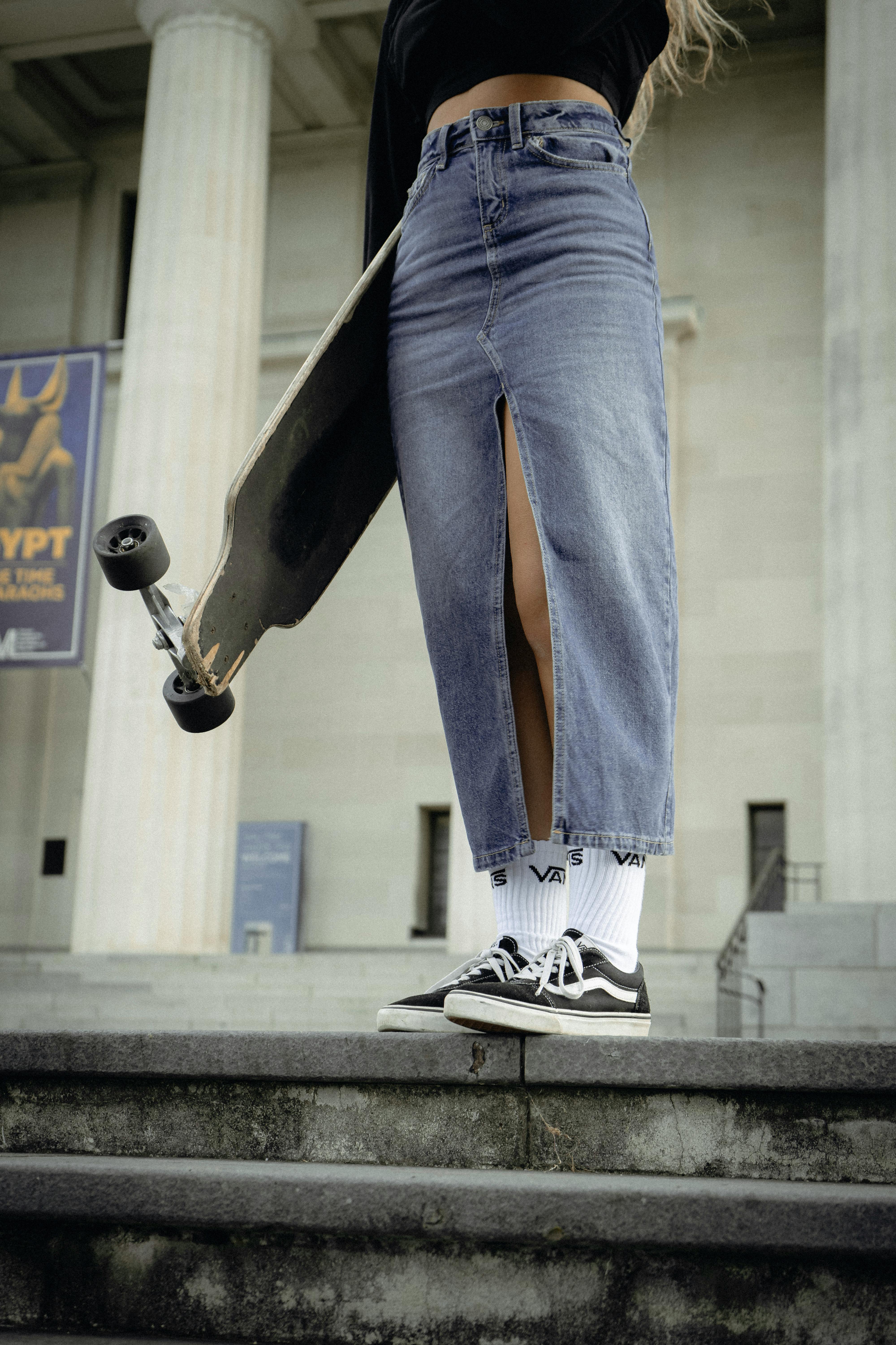 Cropped view of a woman in a denim skirt holding a skateboard, standing on steps in an urban setting.