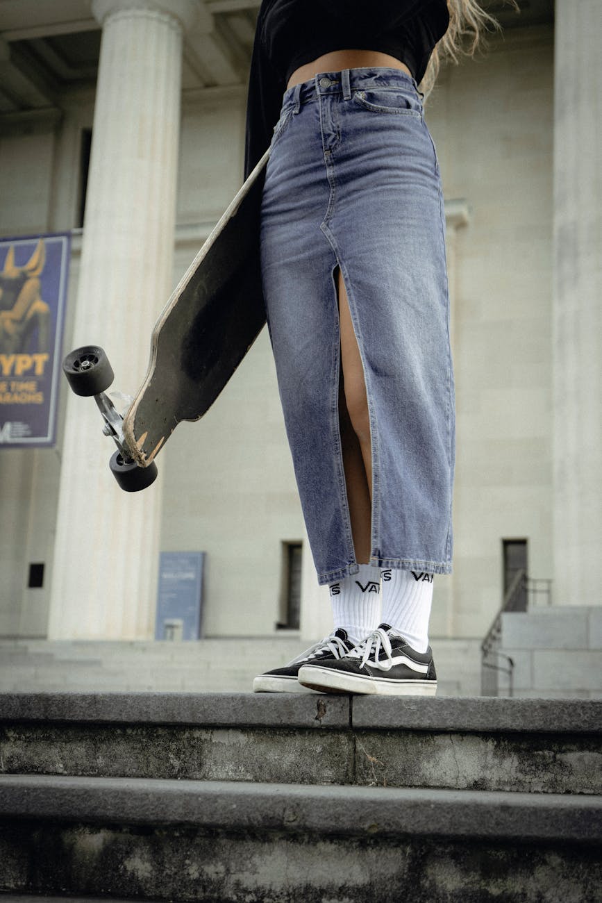Cropped view of a woman in a denim skirt holding a skateboard, standing on steps in an urban setting.