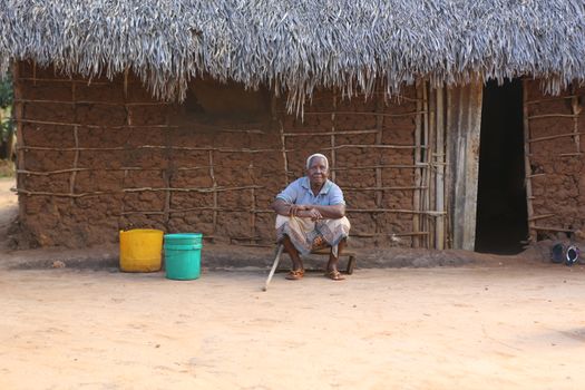 An elderly woman sits in front of a traditional thatched roof hut in a rural village.