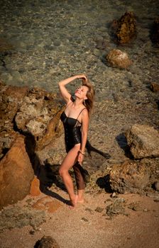 Woman in swimsuit posing elegantly among rocks on a sunny beach day.