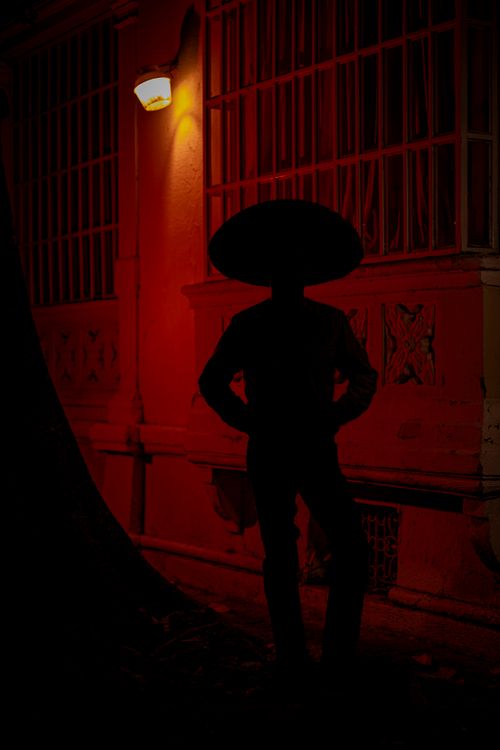 Free A silhouette of a person in a hat stands in dramatic lighting beneath a streetlamp at night. Stock Photo