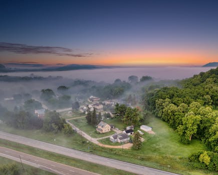 Bird's eye view of a mist-covered rural village surrounded by lush greenery at sunrise.