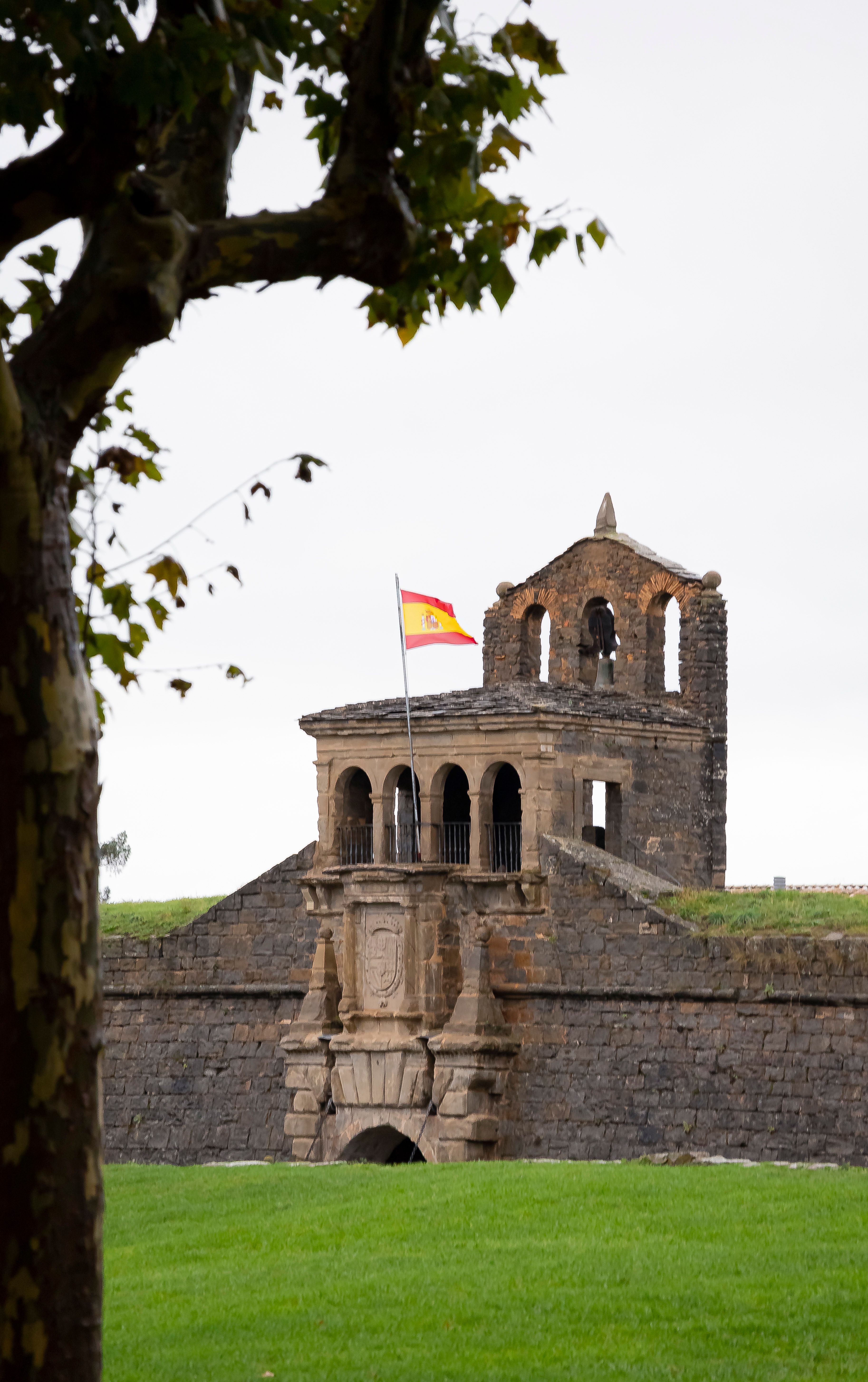 A historic stone fortress featuring a Spanish flag, captured with lush greenery and autumn leaves.