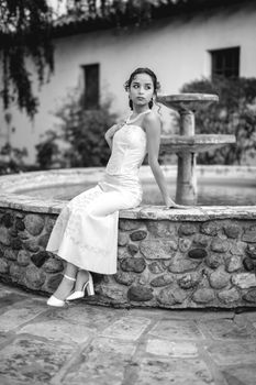 A stylish woman in a white dress poses elegantly by a stone fountain in a black and white photograph.