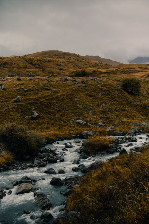 Free A tranquil river flows through a rocky valley under cloudy skies in autumn scenery. Stock Photo