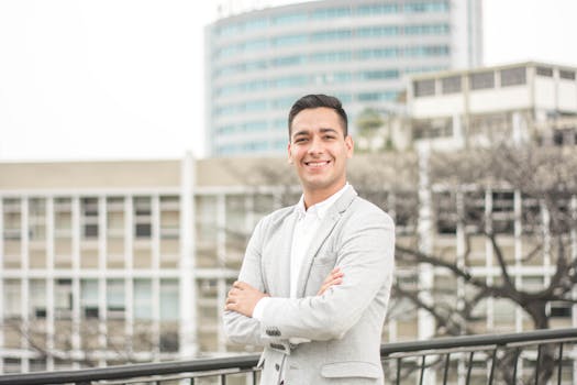 Confident man in a light suit smiling in a city environment with modern buildings.