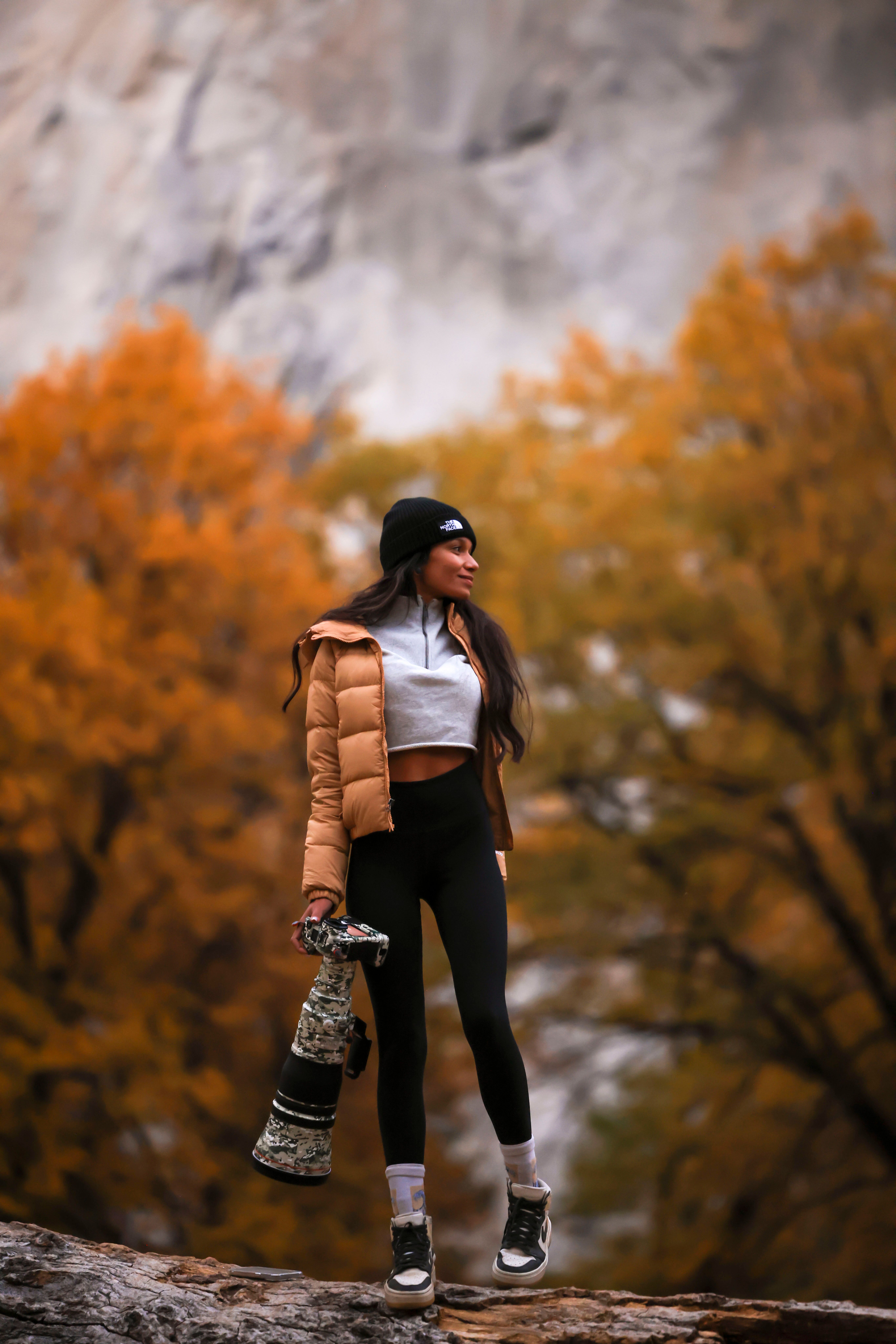 Woman in a brown jacket holding a camera with fall colors in the background.