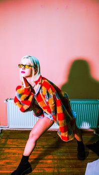 Stylish woman with sunglasses and colorful coat posing against a pink wall indoors.