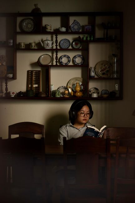 A teenager reads a book wearing headphones in a dimly lit kitchen filled with vintage tableware.