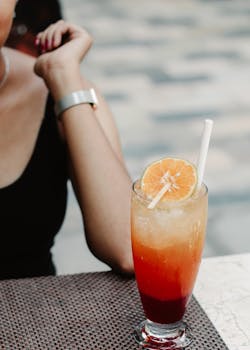 Vibrant orange cocktail with straw and ice on a woven mat with a woman's arm in view.