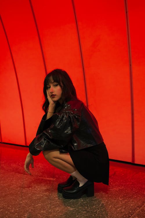 Young Brunette Woman in Leather Jacket and Skirt Crouching by Red Wall