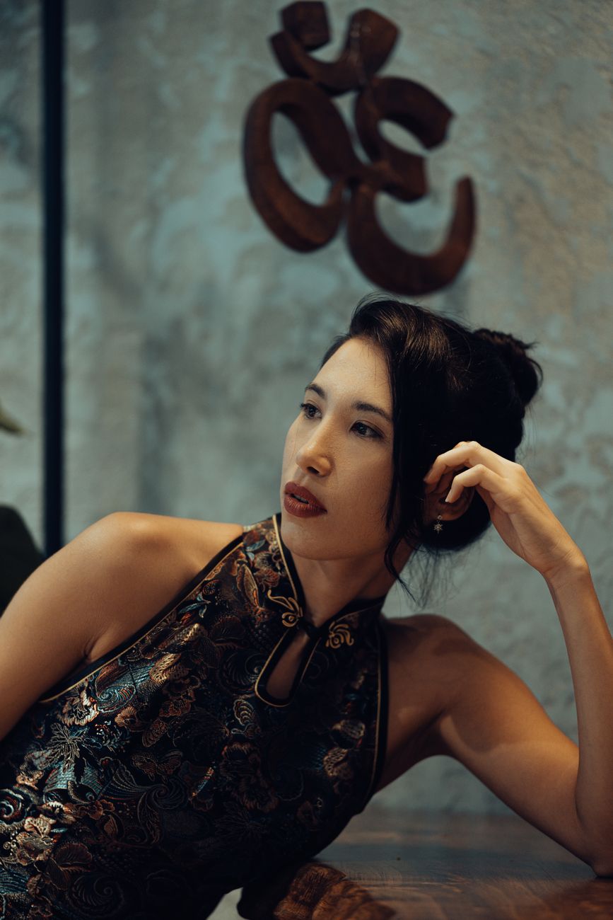A stylish woman in traditional clothing poses elegantly below an Om symbol indoors.