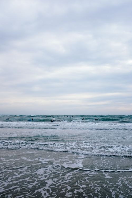 Free Peaceful beach scene with gentle waves, cloudy sky, and distant figures enjoying the sea. Stock Photo