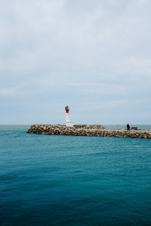 Free A solitary red and white lighthouse stands on a rocky pier by the sea on a cloudy day. Stock Photo