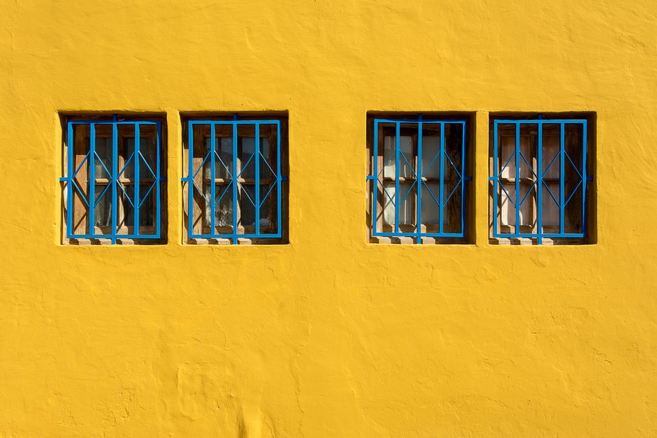 Bright yellow exterior wall featuring four blue-barred windows under sunlight.