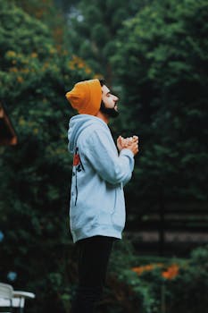 一名男子头戴无檐帽、身穿连帽衫，在郁郁葱葱的森林中虔诚祈祷