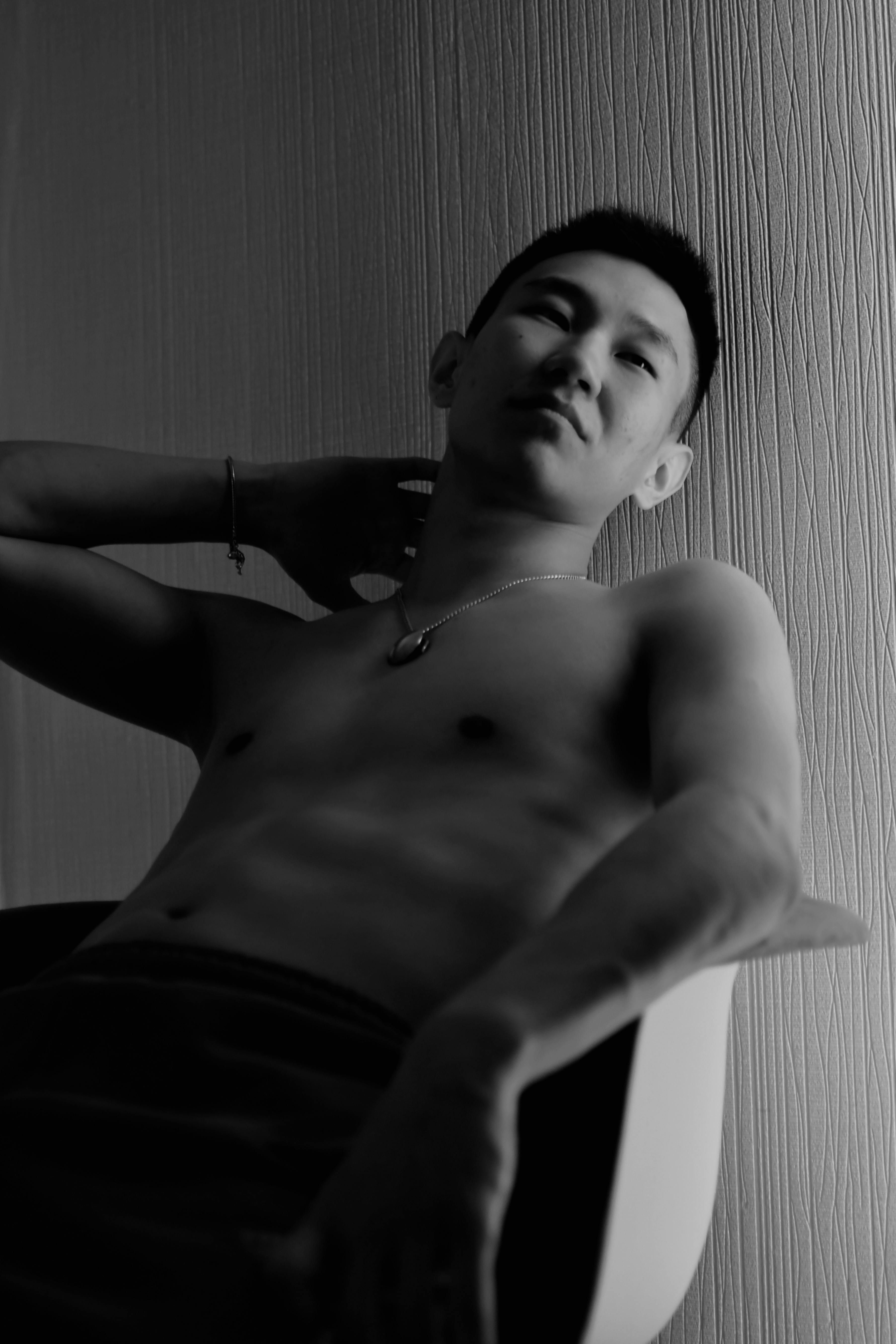 Dramatic black and white portrait of a shirtless Asian man sitting and posing in a chair.