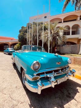 A classic blue convertible car parked in front of a tropical resort under bright sunlight.