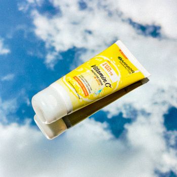 Tube of Vitamin C skincare cream reflecting in mirror against a bright sky.
