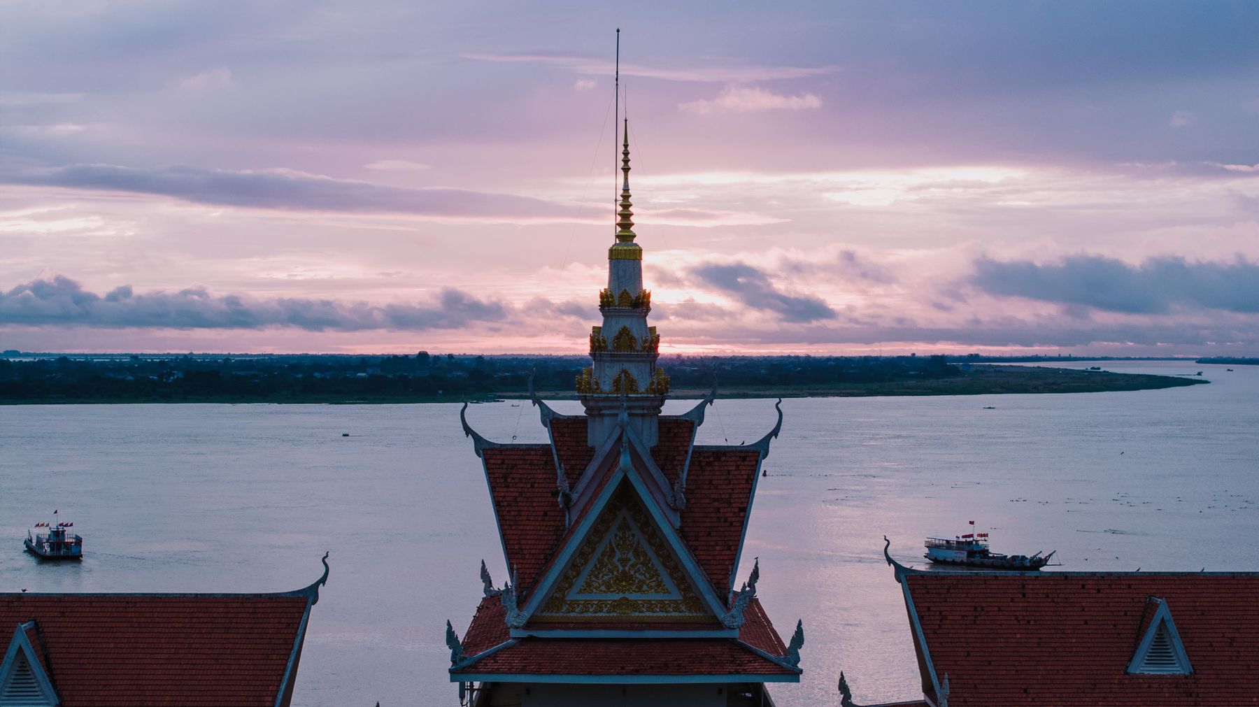 Passenger boats moving below an ornate Khmer temple spire on the Mekong at sunset