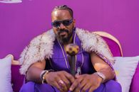 Confident man wearing purple and fur cape, sitting with sunglasses in studio.