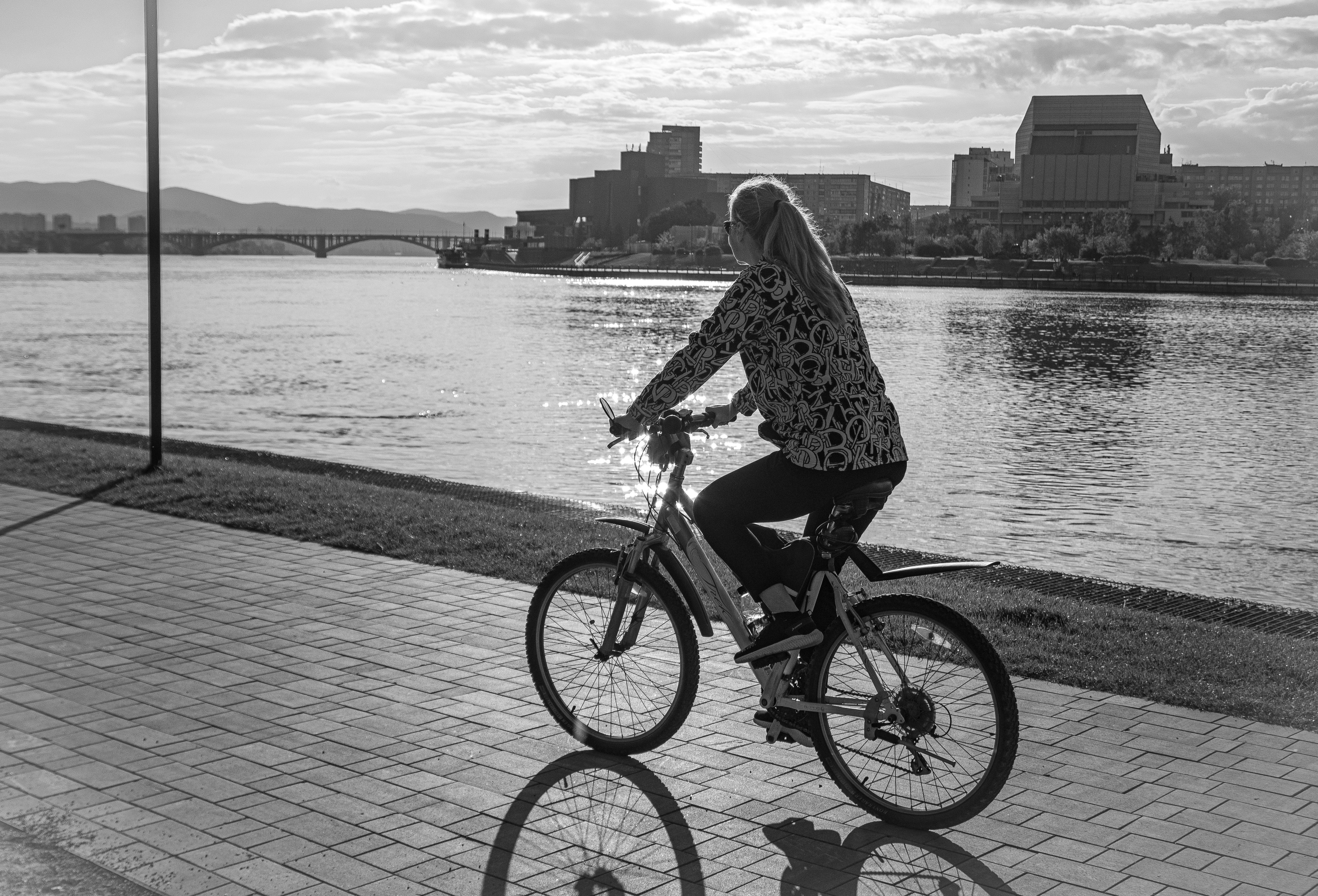 A woman rides a bicycle on a riverside path, with cityscape and bridge in the background.