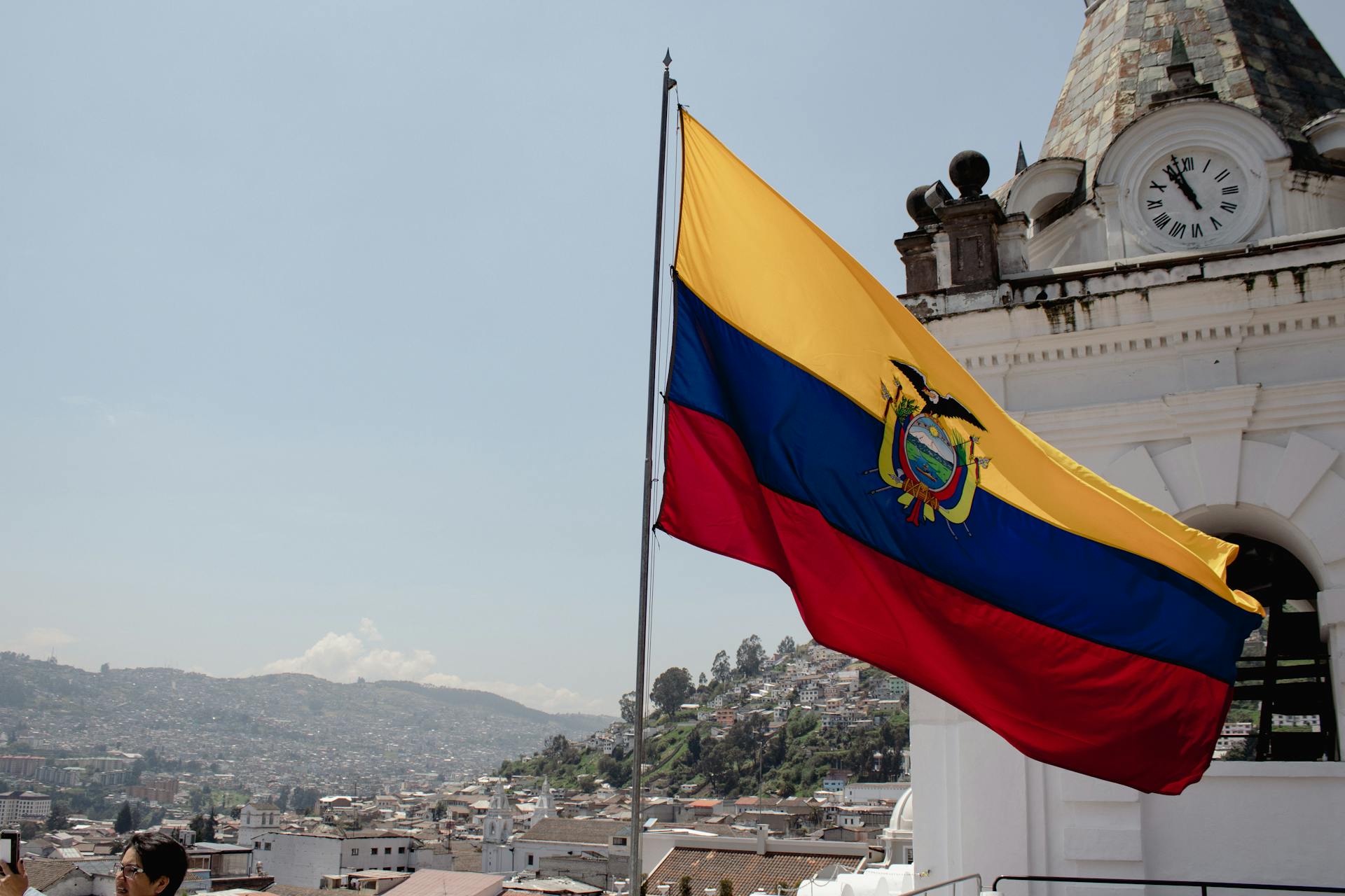 A vibrant Ecuadorian flag waves over the cityscape of Quito with a historic clock tower.