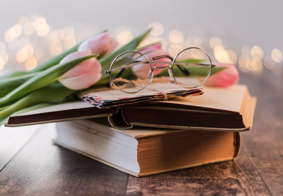 Artistic composition of vintage books, eyeglasses, and pink tulips on a wooden table with bokeh lights.