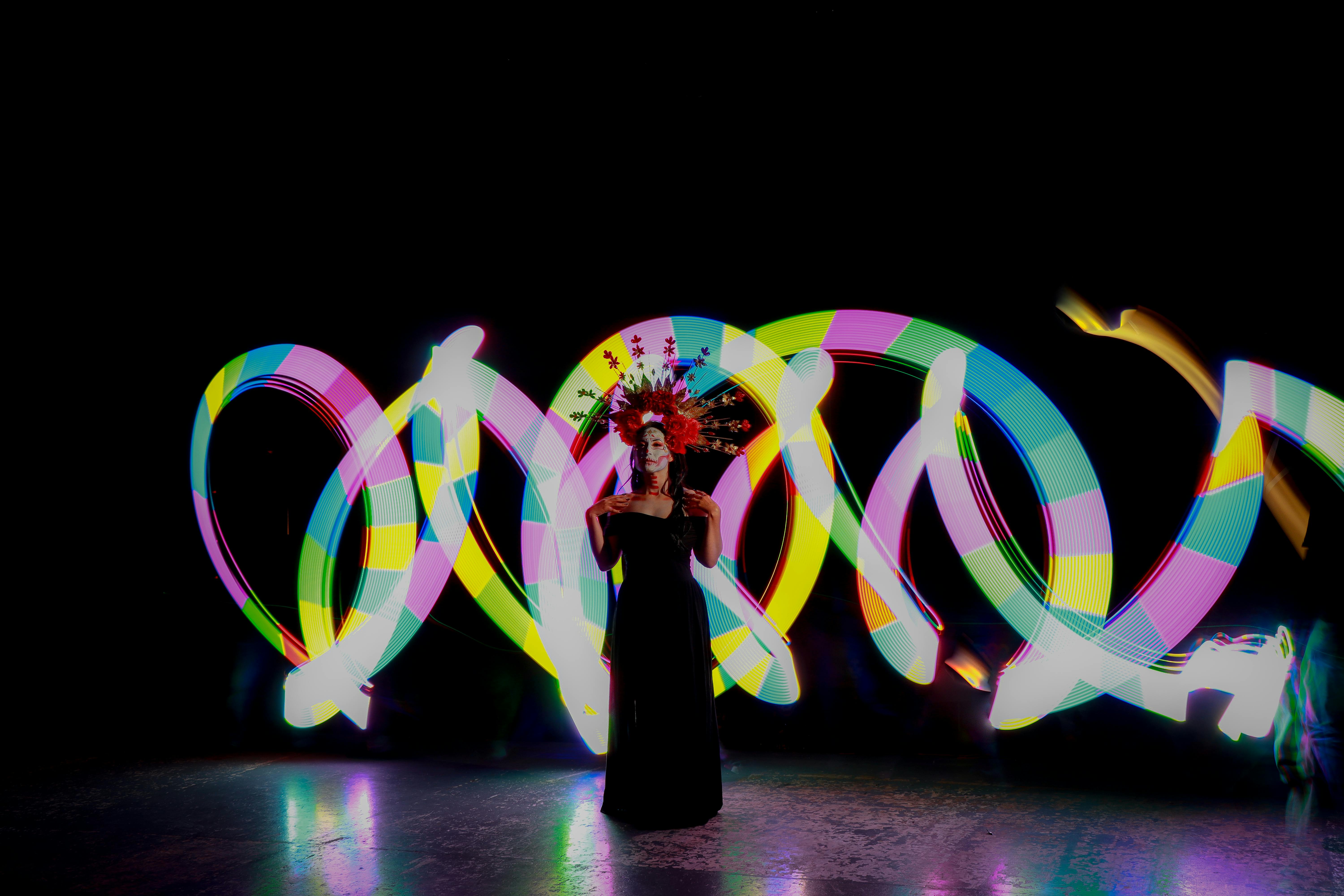 A woman in traditional Dia de Muertos attire stands amidst colorful light trails, showcasing Mexican culture.