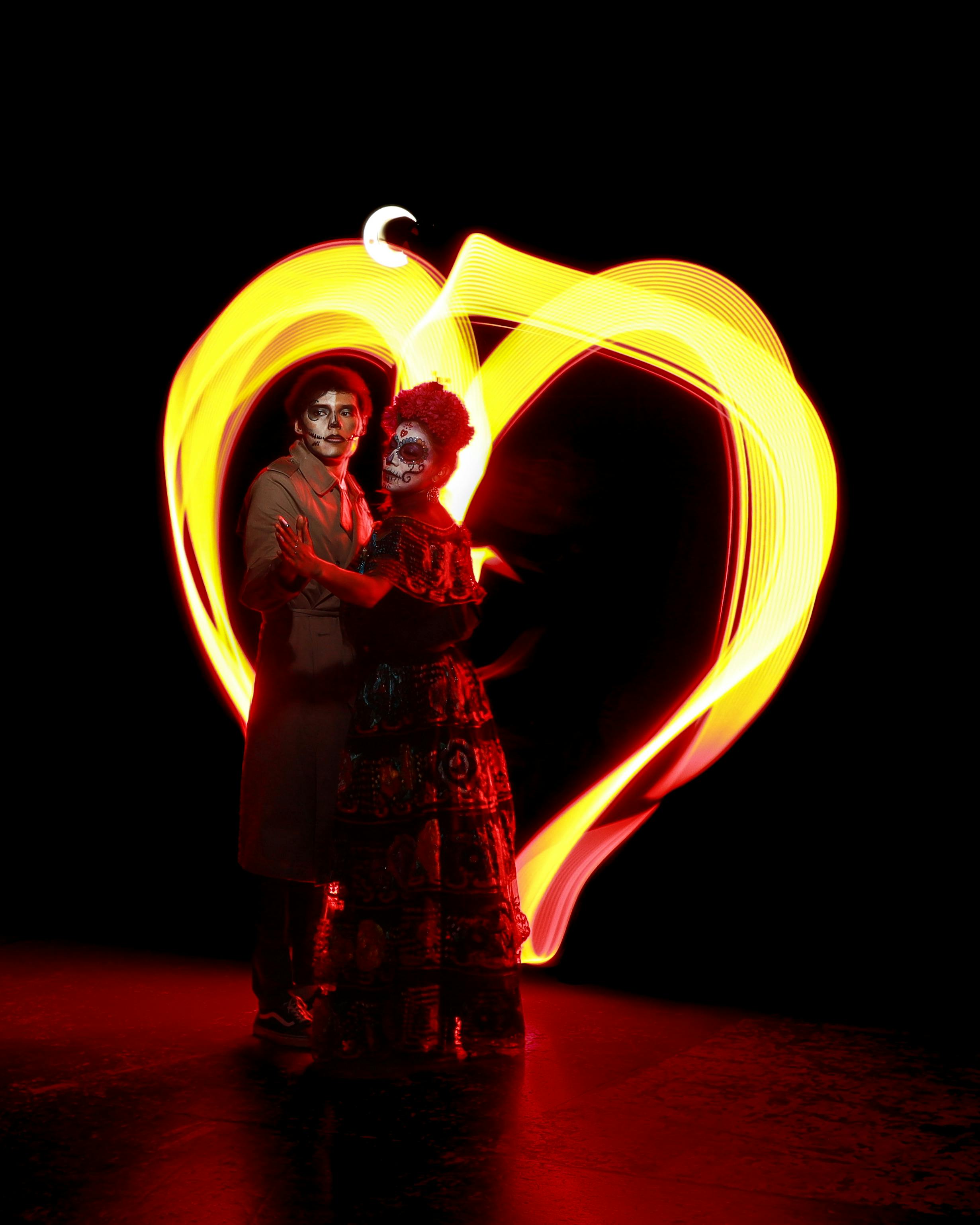 Couple in Día de Muertos attire illuminated by vibrant light painting heart shape.