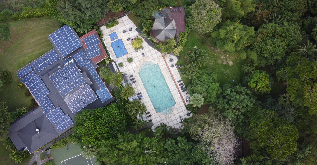 Aerial view of a luxury villa with solar panels and a pool in vibrant green surroundings.