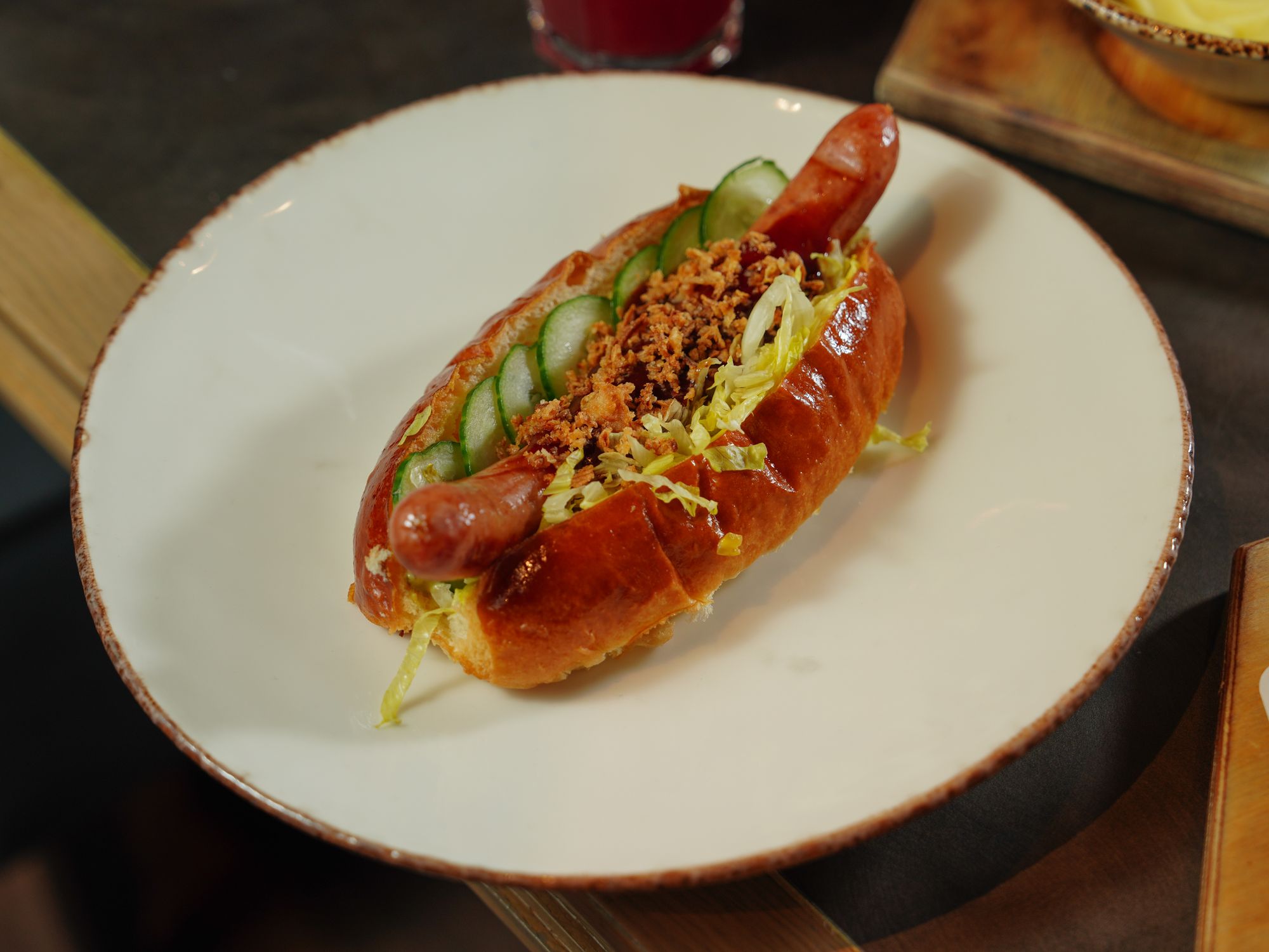 A craft link in a toasted bun, dressed with sliced cucumber, shredded lettuce and crisp fried onions