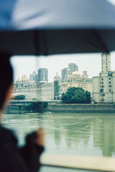 A person holds an umbrella overlooking a river with a cityscape in the background, capturing a rainy urban scene.