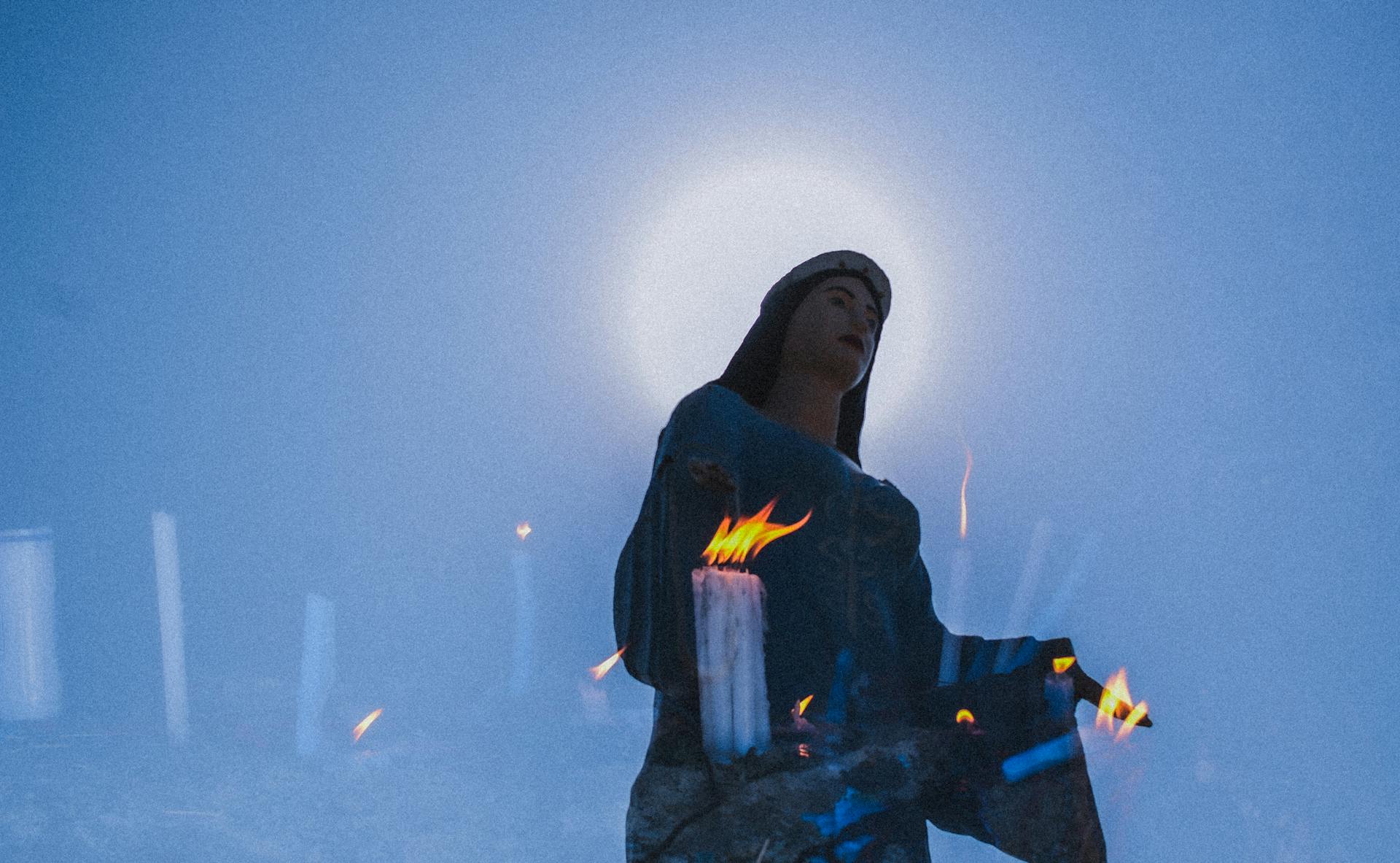 Lit Candles Behind a Glass Reflecting the Statue of the Virgin Mary with a Halo of the Sun Above Head